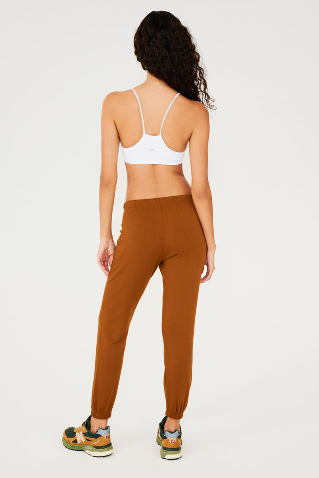 Full back view of woman with dark wavy hair wearing dark orange sweatpant jogger with white drawstring and white bra with racerback spaghetti straps paired with dark green, orange and light green shoes