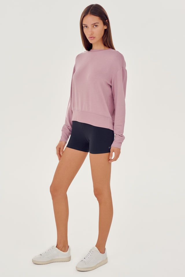 Full front side view of woman with dark brown hair wearing a light pink crewneck sweatshirt and black shorts paired with white shoes