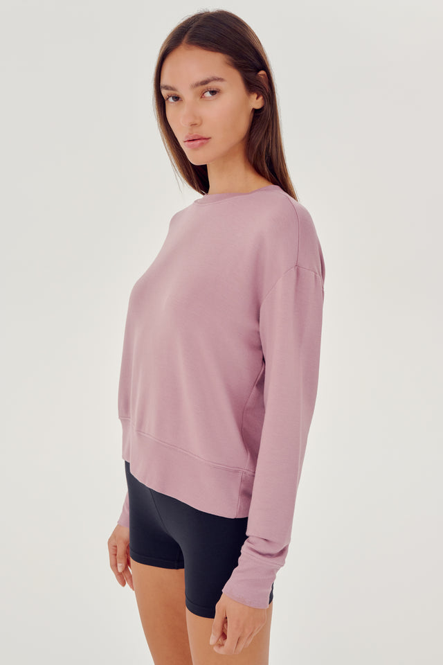 Front side view of woman with dark brown hair wearing a light pink crewneck sweatshirt and black shorts