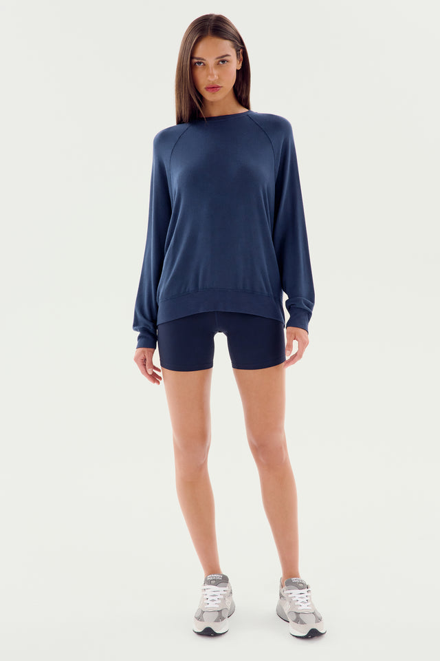 A woman stands wearing an indigo long-sleeve Andie Fleece Sweatshirt by SPLITS59, paired with black shorts and white sneakers, set against a plain white background. MADE IN LOS ANGELES.