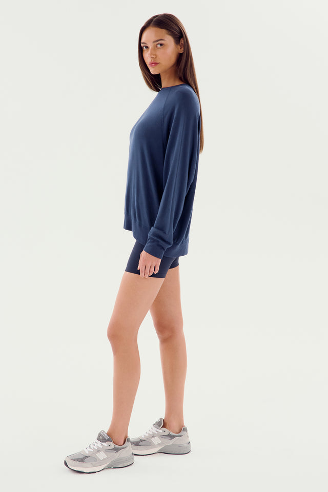 A person standing sideways, wearing a SPLITS59 Andie Fleece Sweatshirt in Indigo, navy shorts, and gray sneakers against a plain white background. The sweatshirt is designed and MADE IN LOS ANGELES.