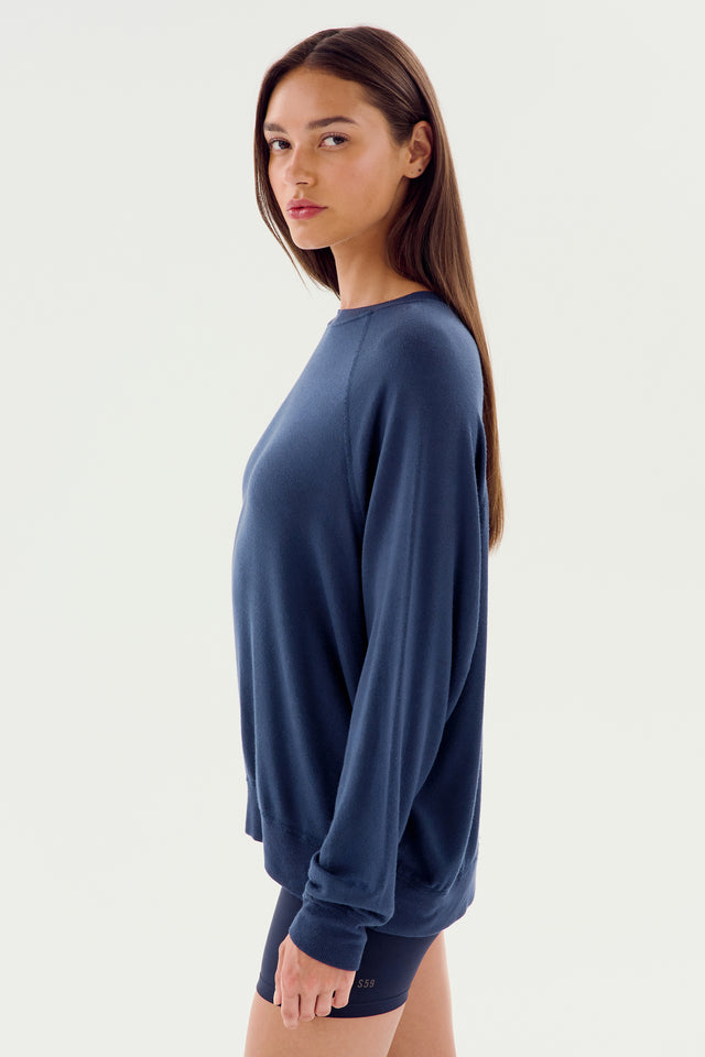 A woman stands sideways, wearing an indigo Andie Fleece Sweatshirt by SPLITS59 and black leggings, gazing at the camera against a plain white background.