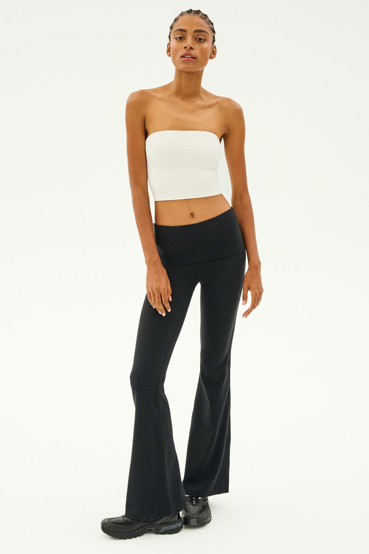 A person stands against a plain background, wearing the SPLITS59 Hazel Airweight Crop Bandeau in white paired with black flared pants made from stretch fabric and completing the look with black sneakers.