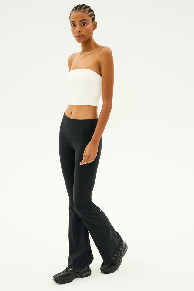 A person stands against a plain background, showcasing the SPLITS59 Hazel Airweight Crop Bandeau in white and black flared pants made from stretch fabric. Black sneakers complete the sleek look.