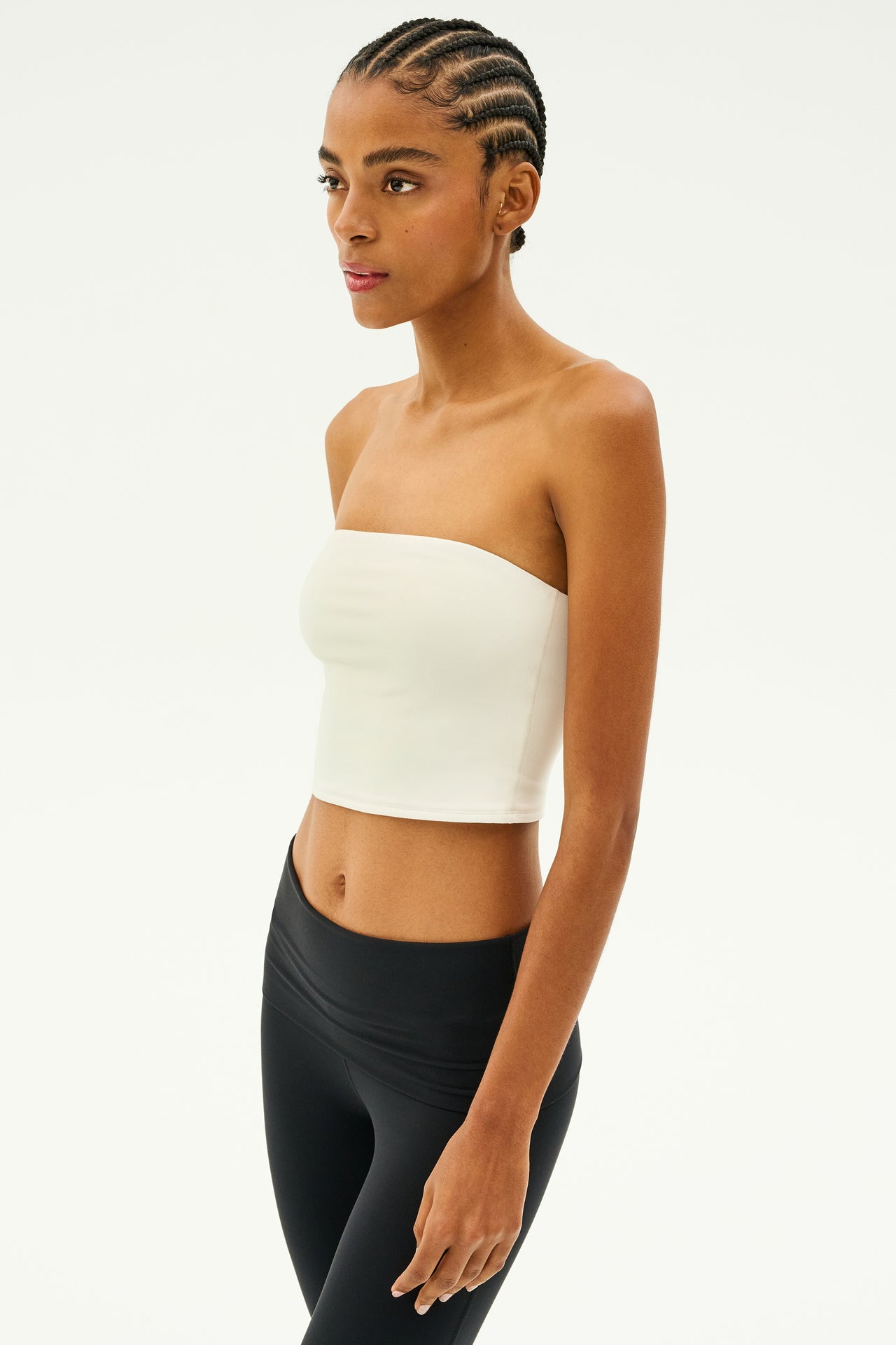 A person with braided hair stands against a plain background, showcasing the support of a white Hazel Airweight Crop Bandeau by SPLITS59, paired with black leggings made from high-quality stretch fabric.