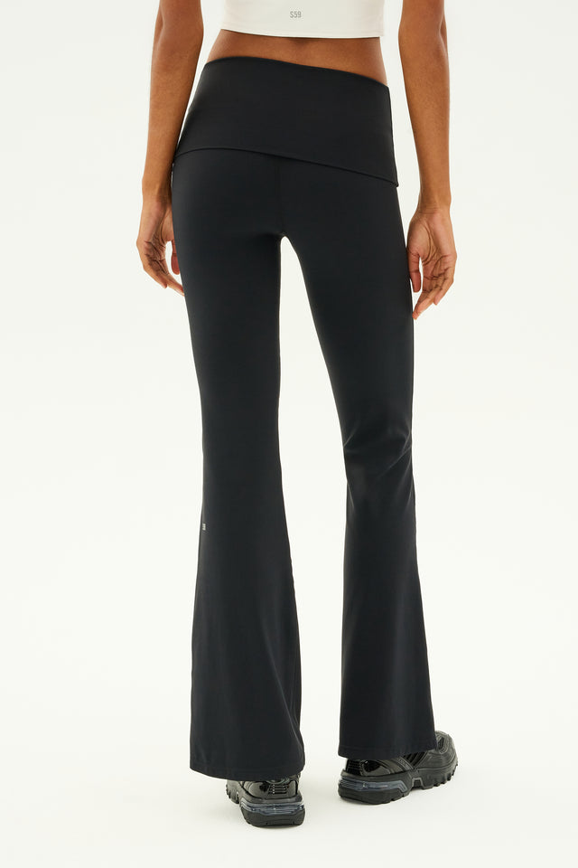 A person in SPLITS59's Raquel Airweight Foldover Flare pants in black and a white top stands with their back to the camera.