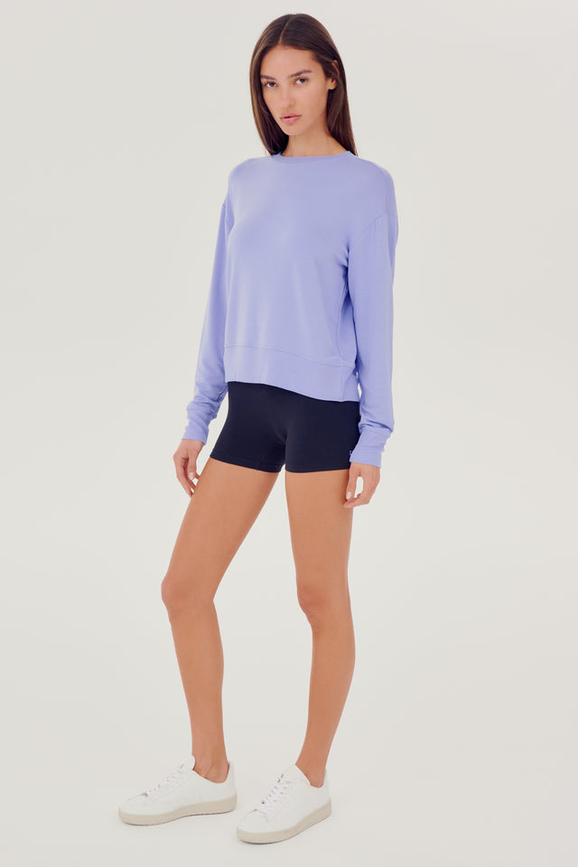 Full front side view of woman with dark brown hair wearing a light purple crewneck sweatshirt and black shorts paired with white shoes