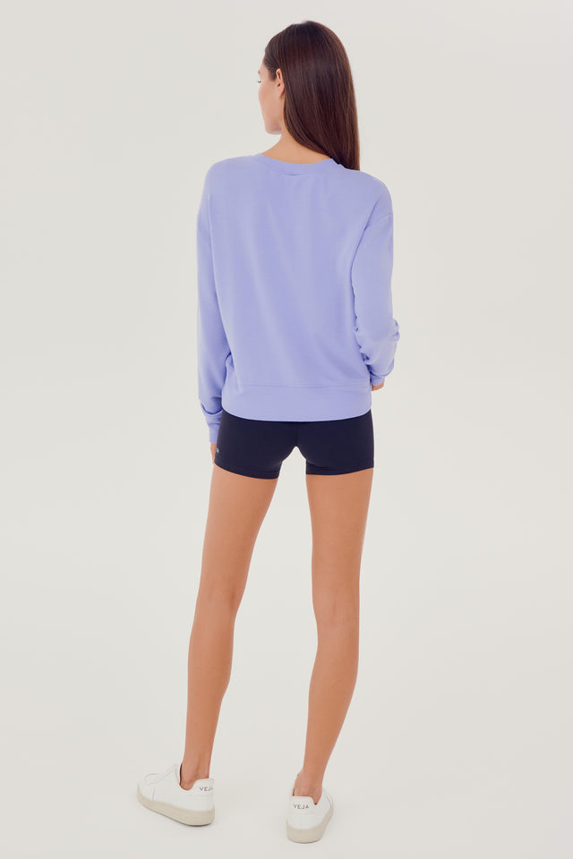 Full back view of woman with dark blonde hair wearing a light purple crewneck sweatshirt and black shorts paired with white shoes