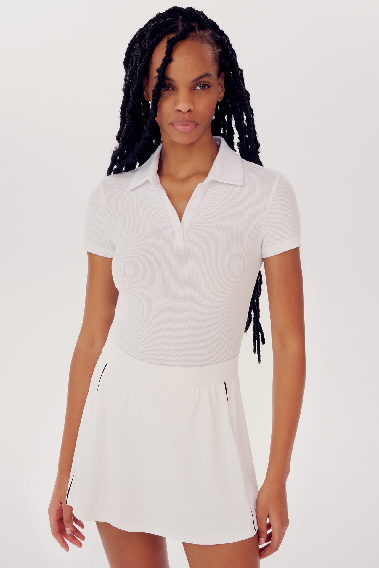 A woman wearing a SPLITS59 Talia Rib Polo in White and skirt, standing against a plain background, looks directly at the camera. Her hair is styled in long black braids for yoga.