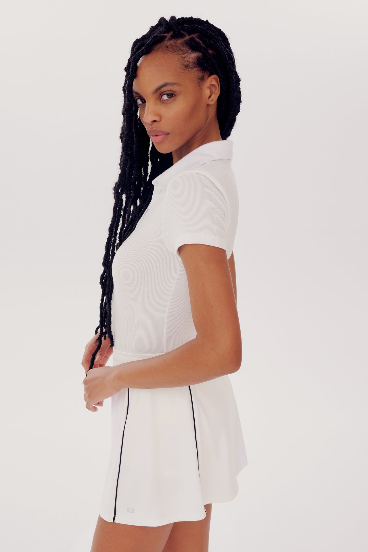 A young woman wearing a SPLITS59 Talia Rib Polo in white, posing side-profile with her hair styled in long braids, ready for a yoga session.