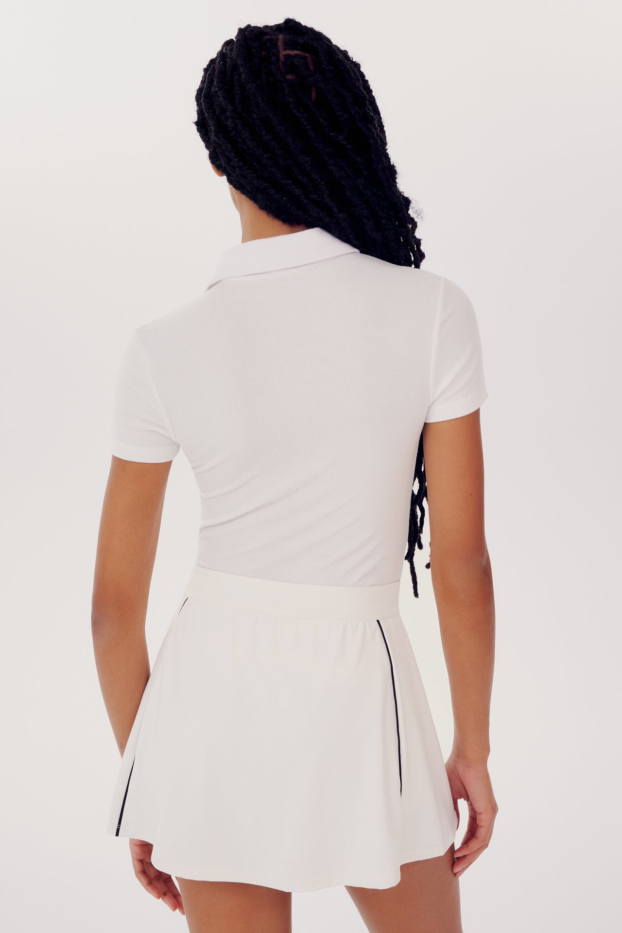 A woman facing away, wearing a SPLITS59 Talia Rib Polo - White and pleated skirt, with braided hair hanging down her back, prepared for a yoga session.
