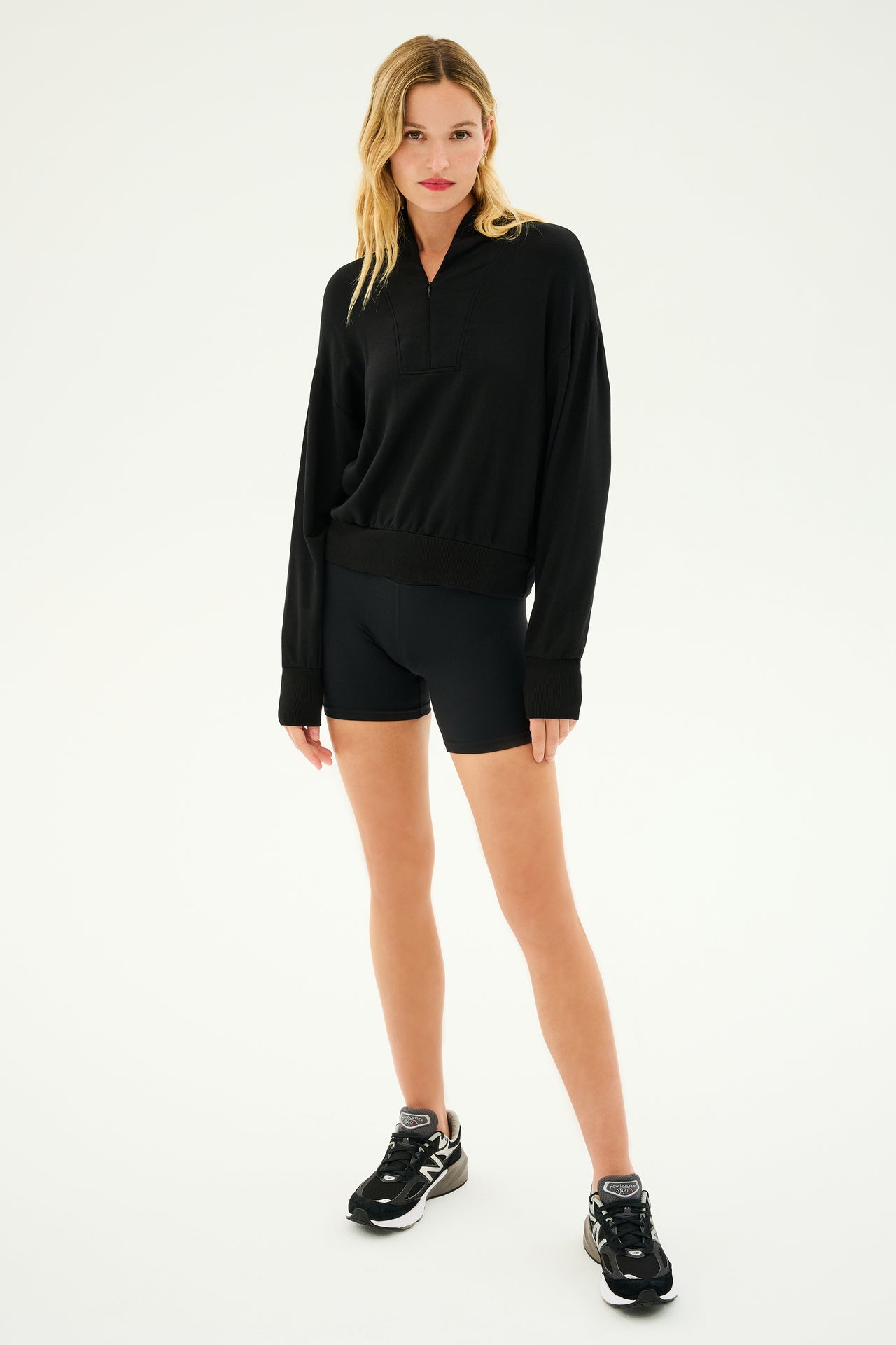 A person stands wearing the Birdie Fleece Half Zip in black from SPLITS59, paired with black shorts made of stretch modal fabric and black sneakers, with arms relaxed at their sides against a plain white background.