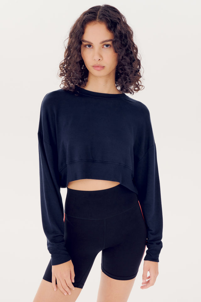 A young woman with curly hair wearing a SPLITS59 Noah Fleece Crop Sweatshirt in Indigo and high-waisted shorts stands against a white background.