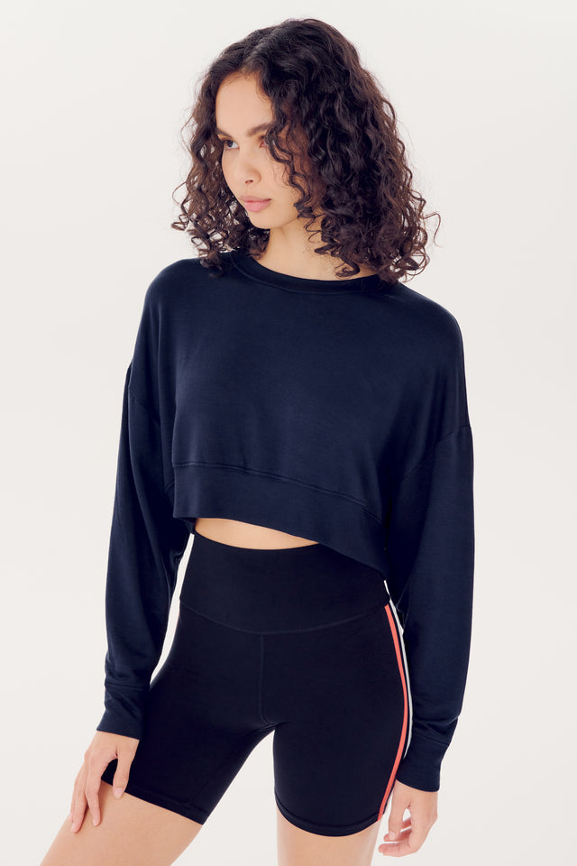 A woman with curly hair wearing a SPLITS59 Noah Fleece Crop Sweatshirt in Indigo and black shorts against a white background.