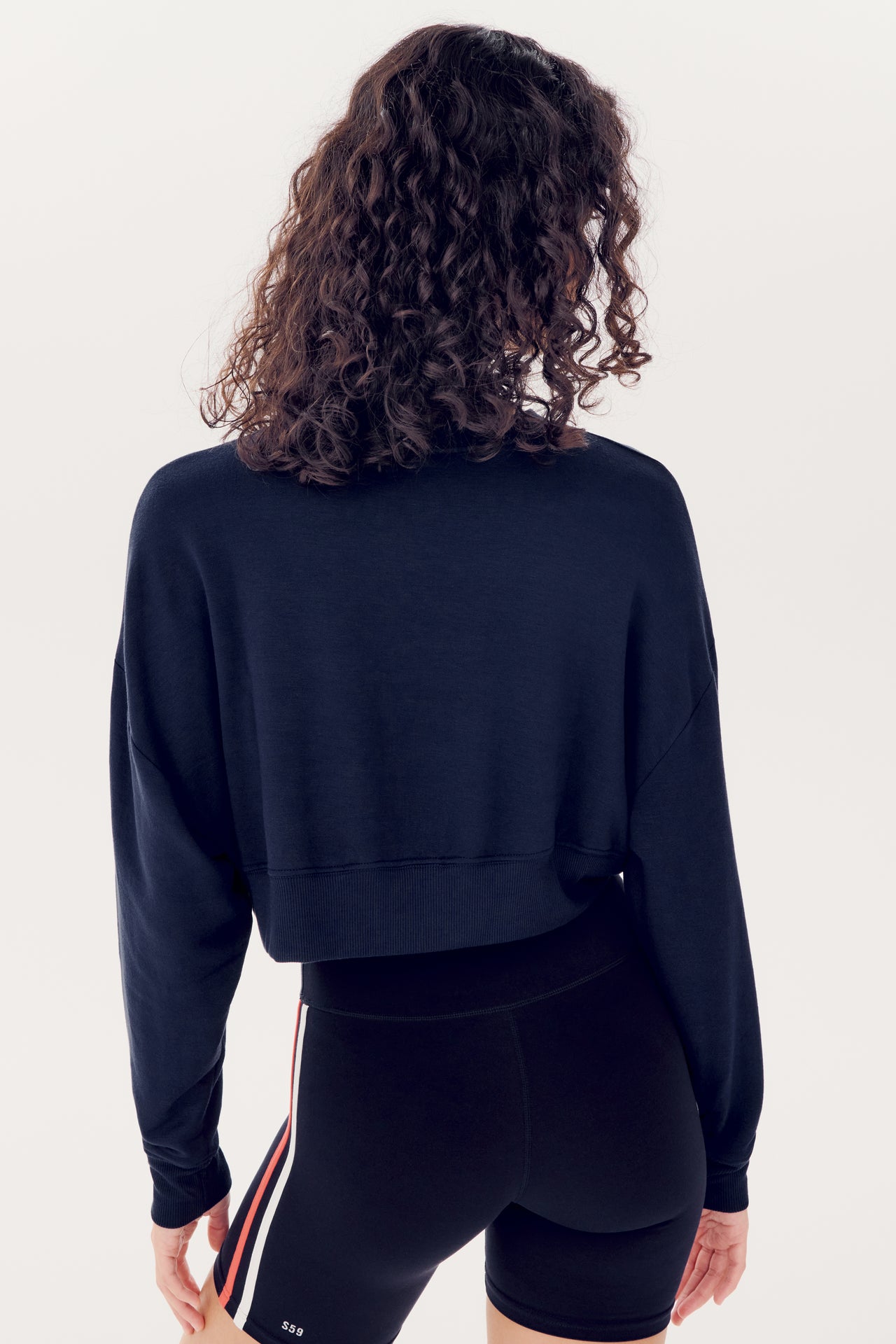 Woman from behind with curly hair, wearing a SPLITS59 Indigo Noah Fleece Crop Sweatshirt and black leggings with a red and white stripe.