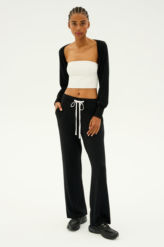 A person poses with hands in their pockets, wearing a white tube top, black shrug, and the Raven Fleece Full Length Sweatpant in Black by SPLITS59. They complete their look with black shoes and braided hair, effortlessly capturing the essence of MADE IN LOS ANGELES style.