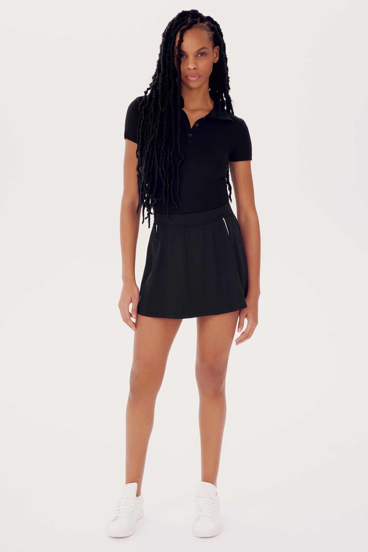 A woman in a black polo shirt and SPLITS59's Venus High Waist Rigor Skort w/Piping - Black stands facing the camera, wearing white sneakers, with long braided hair.