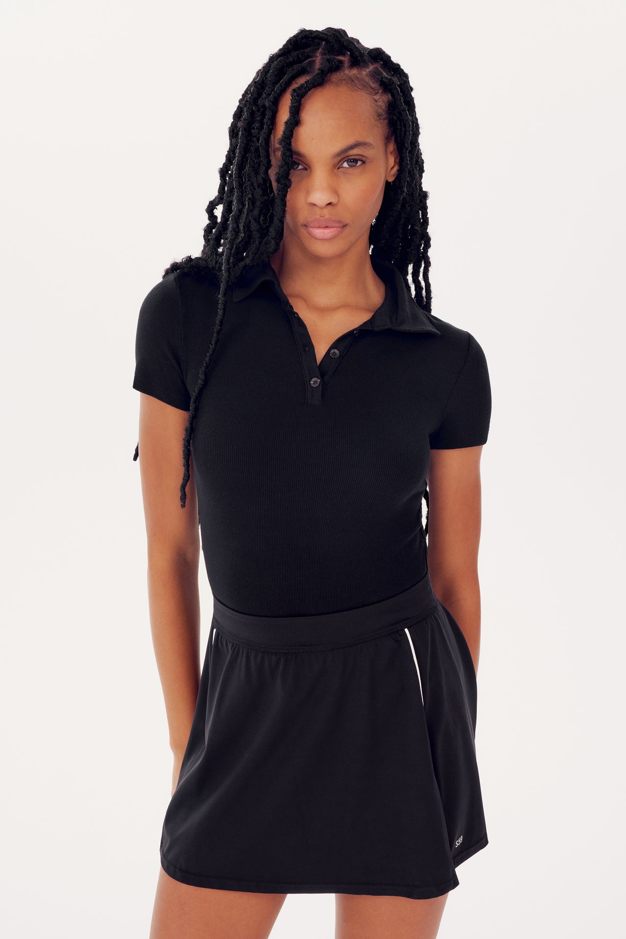 A young woman with braided hair, wearing a SPLITS59 Talia Rib Polo in Black and skirt, stands confidently against a white background.