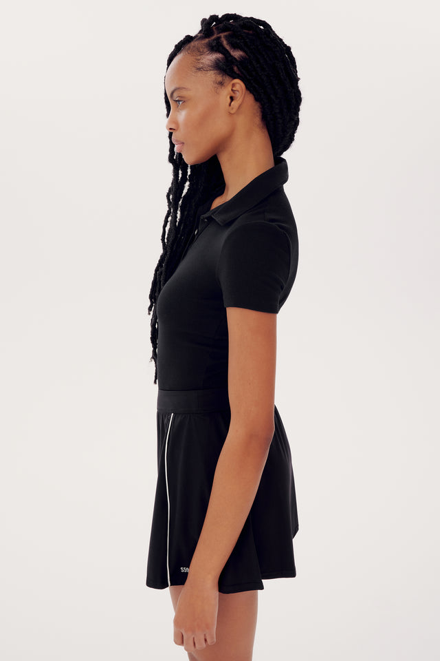 Sentence with replaced product:
Profile view of a black woman with braided hair, wearing a SPLITS59 Talia Rib Polo - Black and a pleated skirt, standing against a white background.