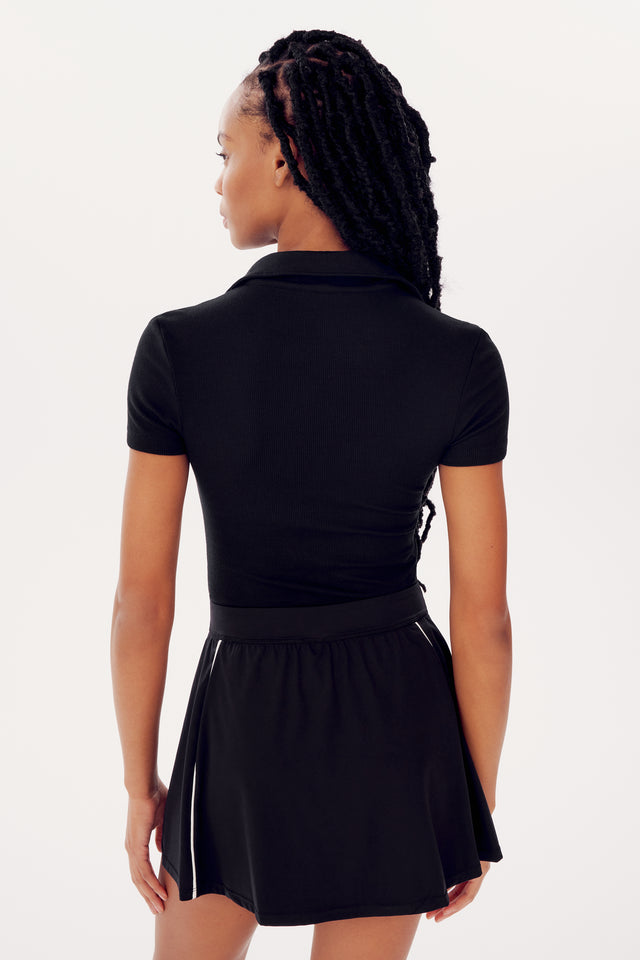 Rear view of a woman with braided hair wearing a SPLITS59 Talia Rib Polo - Black and a black skirt against a white background.