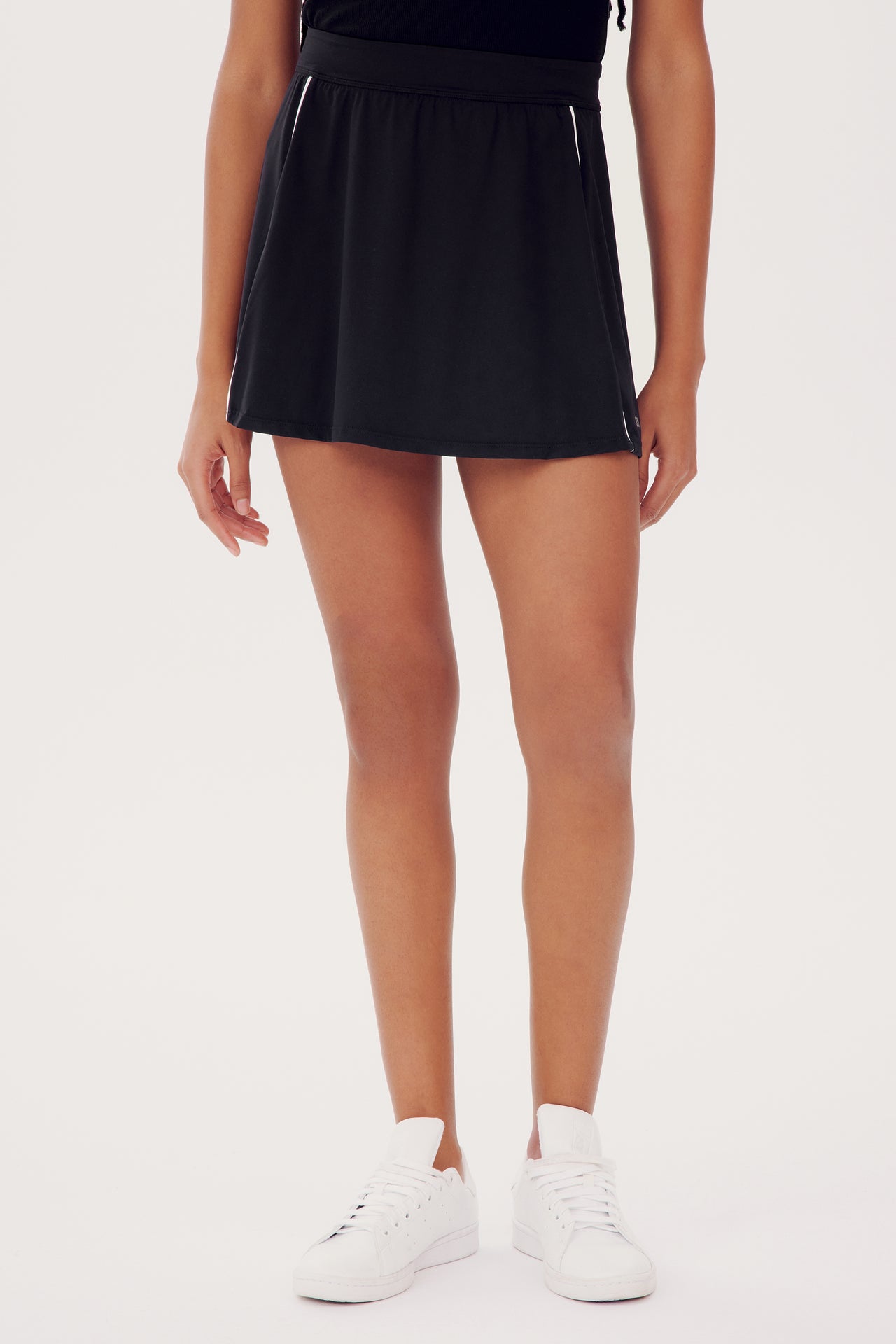 A person wearing a SPLITS59 Venus High Waist Rigor Skort w/Piping in Black and white sneakers stands with hands on hips, cropped at the waist.
