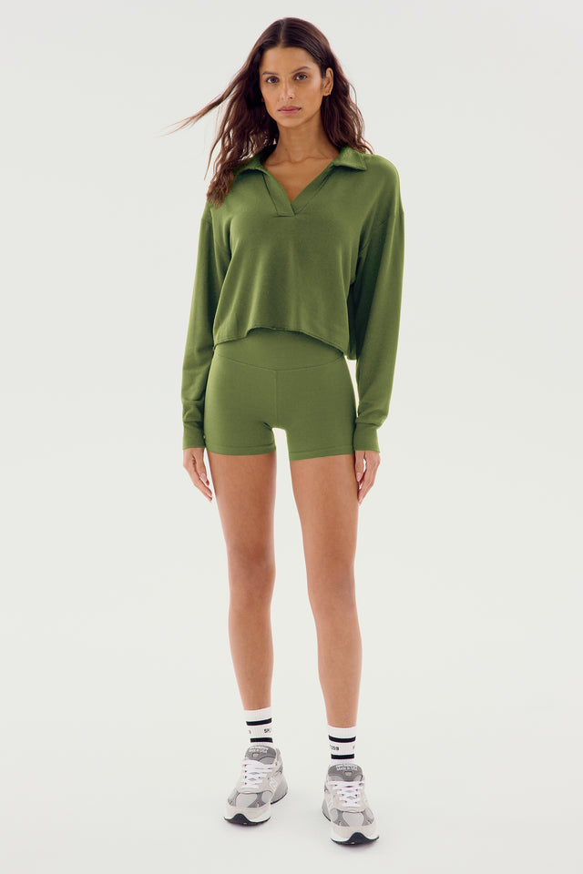 A woman stands against a plain background, wearing the Joan Fleece Polo Sweatshirt in Olive from SPLITS59 and green spandex shorts, complemented by white crew socks and gray sneakers. Her hair is down, and she looks directly at the camera.