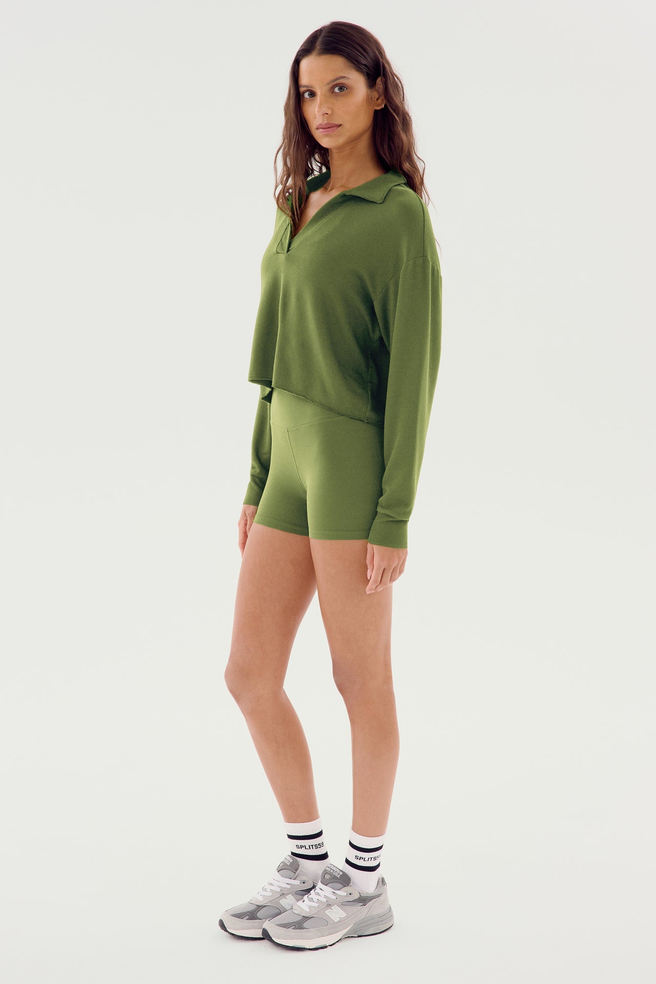 A person stands wearing the Joan Fleece Polo Sweatshirt in Olive and matching green shorts crafted from soft modal fabric, along with gray sneakers and white socks, against a plain white background.