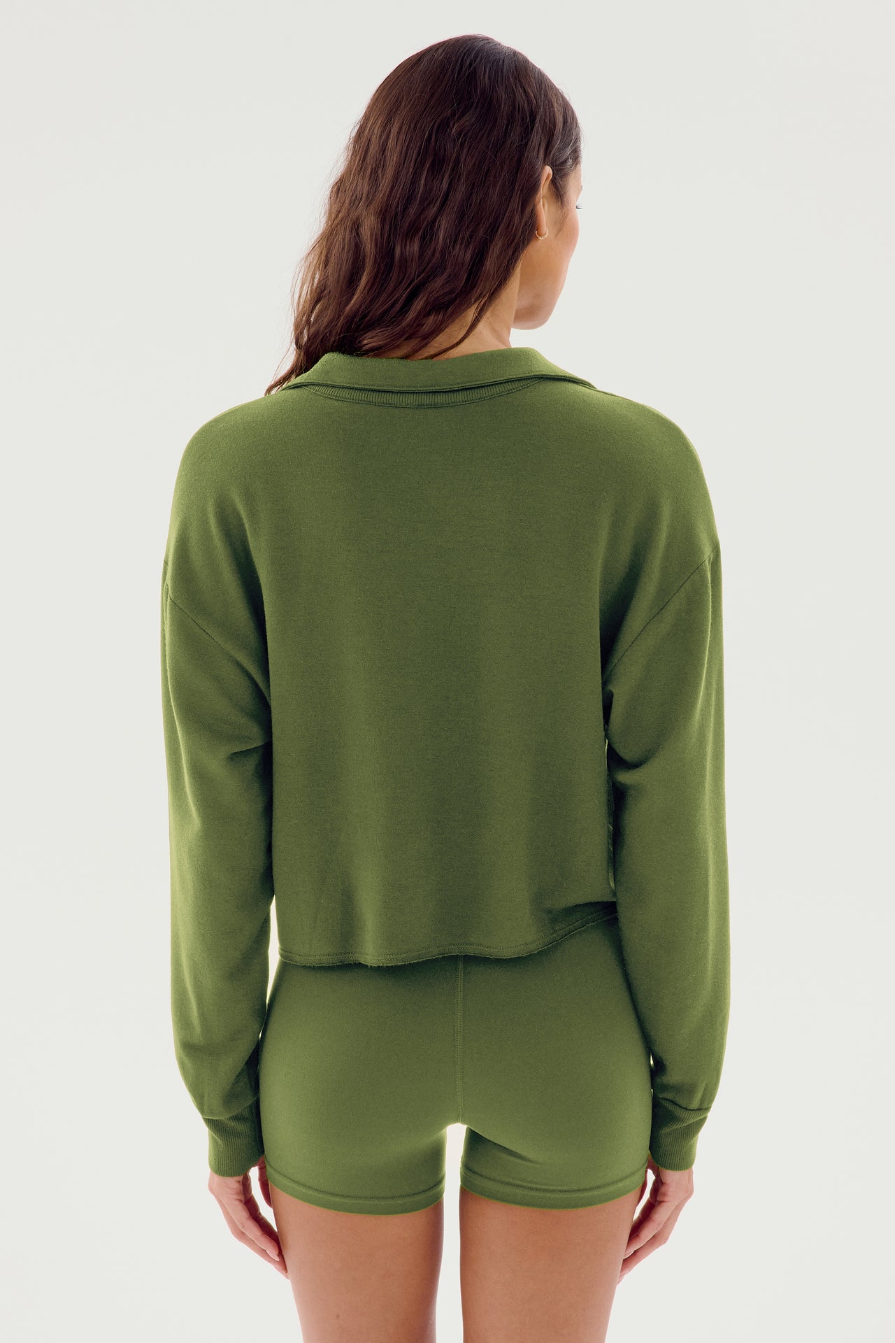 A person with long hair wearing a Joan Fleece Polo Sweatshirt - Olive from SPLITS59 and matching spandex shorts is standing with their back to the camera.