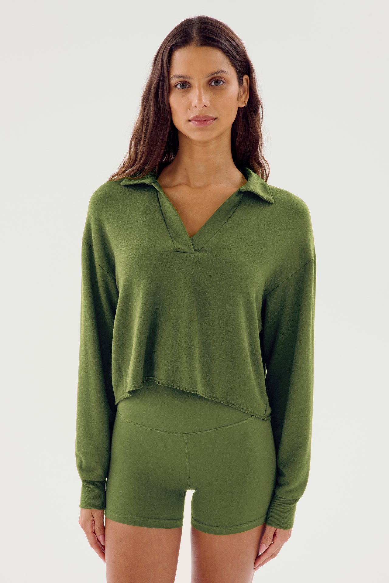 A person with long dark hair is wearing the Joan Fleece Polo Sweatshirt in olive by SPLITS59, paired with green shorts made from 95% modal and 5% spandex, standing against a plain white background.