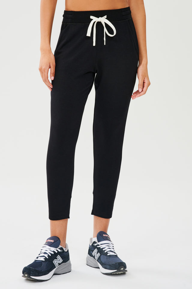 Front view of woman wearing a black sweatpant with tapered leg and above ankle length with white drawstring and side hip pockets. Paired with dark blue and white shoes.