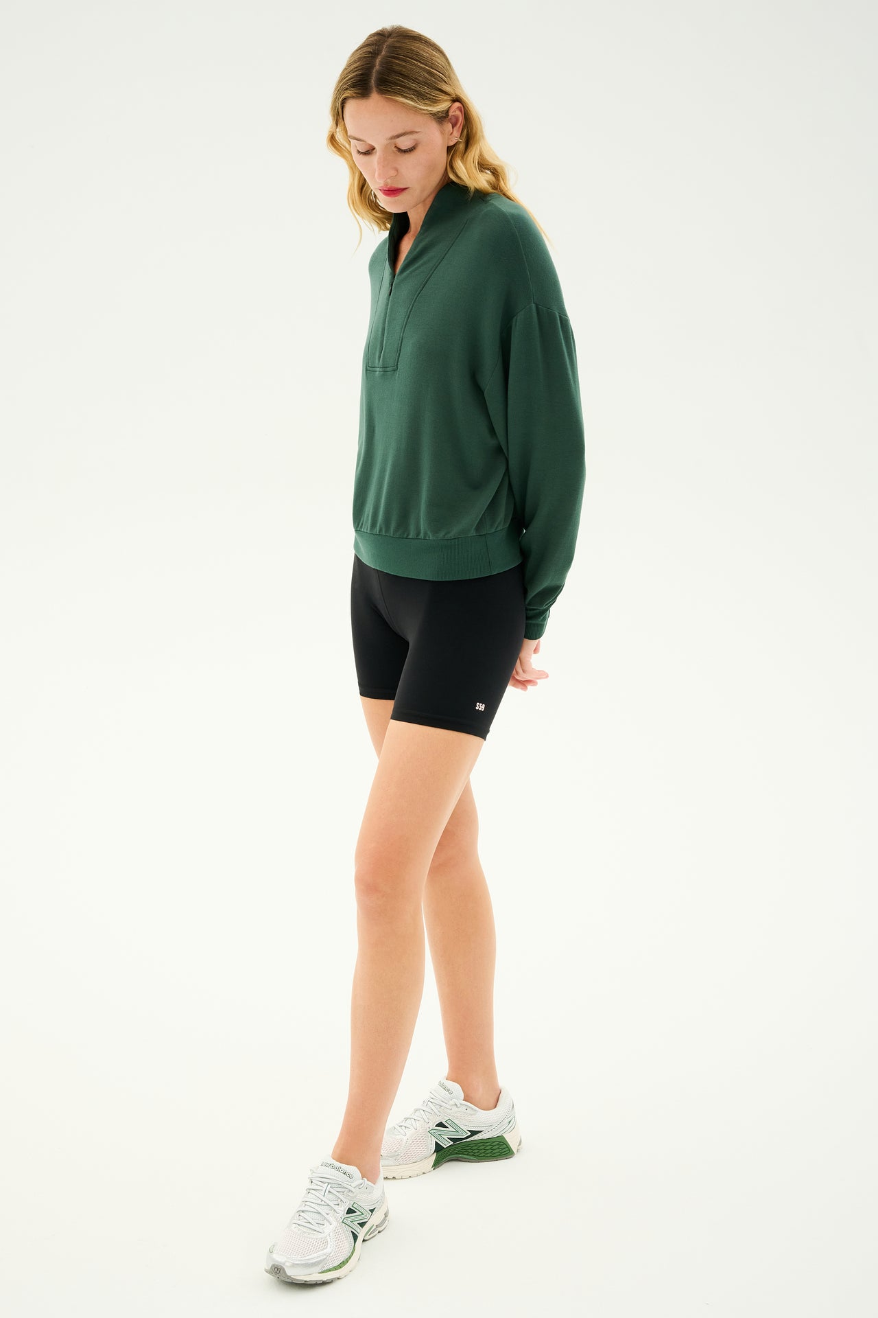 A person stands against a plain backdrop, wearing the cozy Birdie Fleece Half Zip - Military from SPLITS59 made of stretch modal fabric, black shorts, and white sneakers, looking downwards.