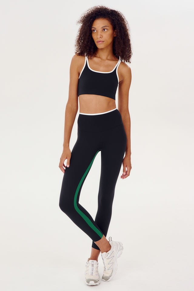 The model is wearing a black and green high-waist sports bra and leggings from SPLITS59's Easton Rigor High Waist 7/8 in Black/White.