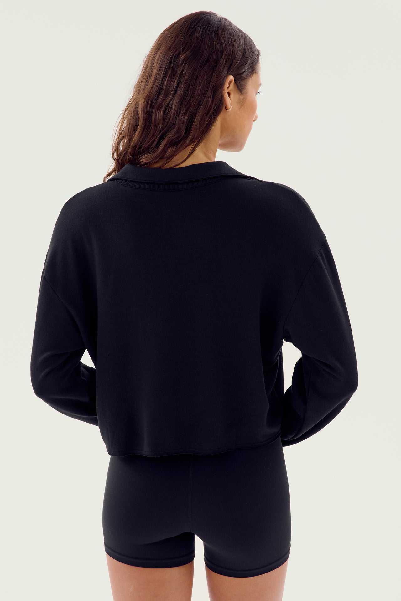 A woman wearing the Joan Fleece Polo Sweatshirt in black by SPLITS59 and black shorts made from high-elasticity modal fabric, faces away from the camera against a plain, light-colored background.