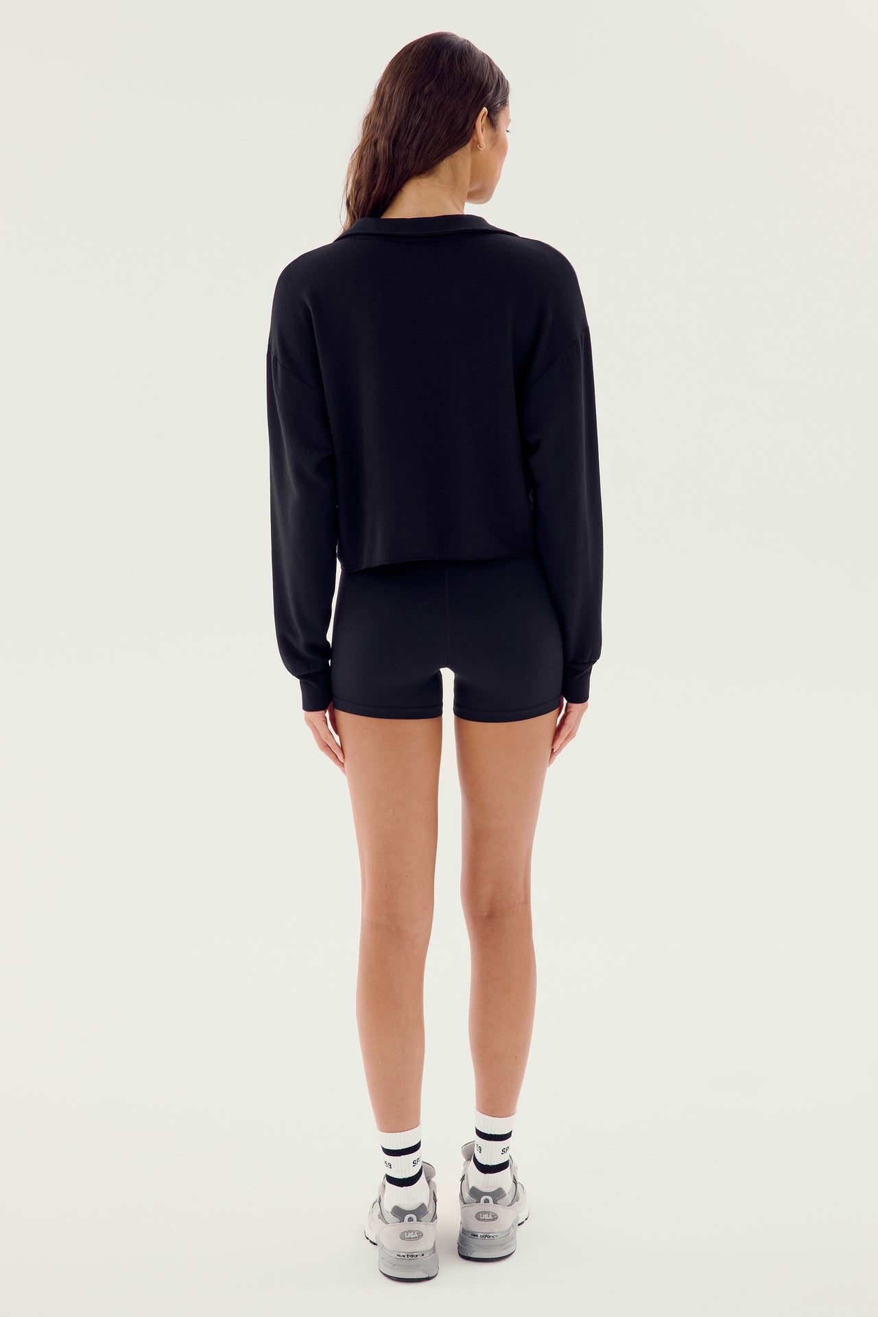 A person with long hair is standing with their back facing the camera, wearing a Joan Fleece Polo Sweatshirt by SPLITS59 in black, black spandex blend shorts, white socks, and grey sneakers against a plain light-colored background.