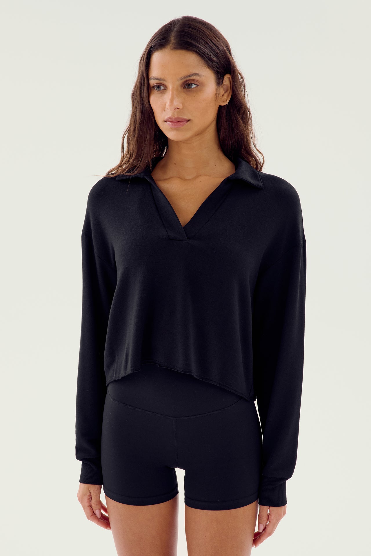 A woman wearing the Joan Fleece Polo Sweatshirt in black from SPLITS59 and black high-waisted shorts made from high elasticity modal fabric is standing against a plain background.