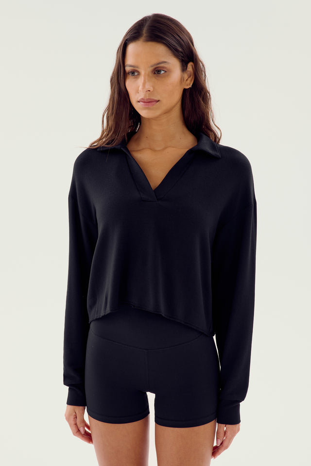 A woman wearing the Joan Fleece Polo Sweatshirt in black from SPLITS59 and black high-waisted shorts made from high elasticity modal fabric is standing against a plain background.