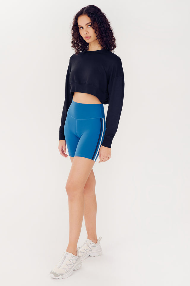 A woman in a SPLITS59 Noah Fleece Crop Sweatshirt in black and blue sports shorts with white stripe, standing against a plain white background.