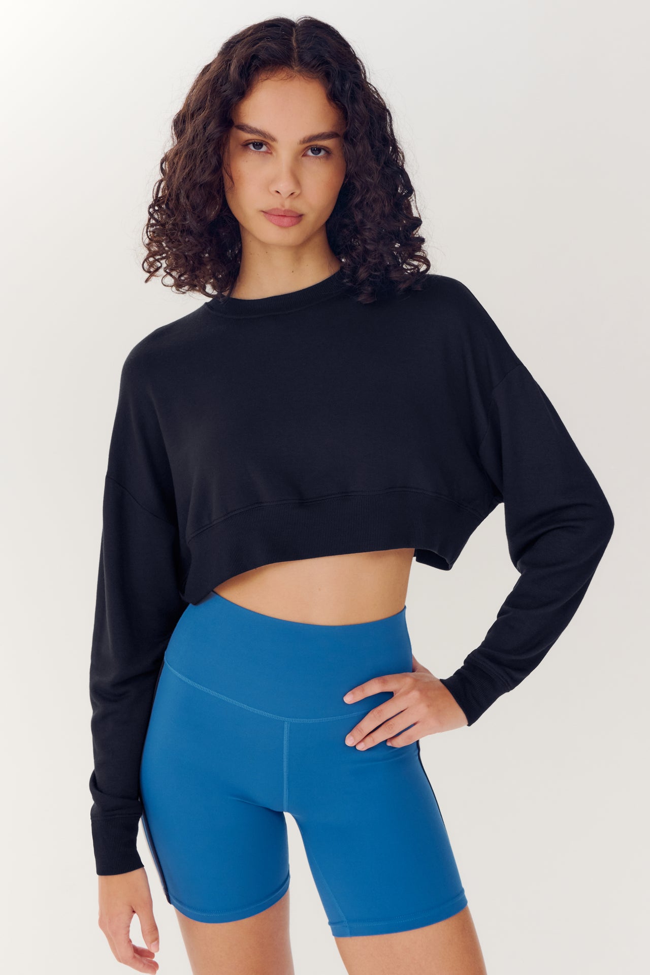 Woman wearing a SPLITS59 Noah Fleece Crop Sweatshirt in Black and blue athletic shorts, standing confidently against a white background.