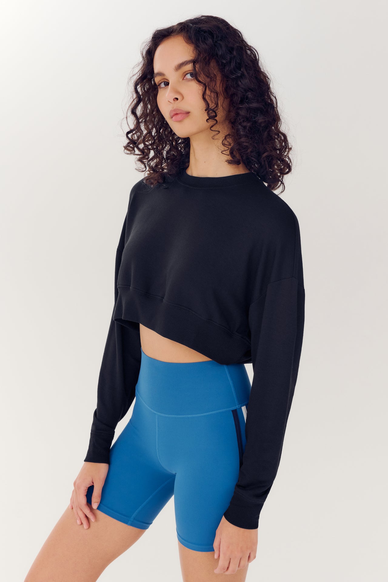 A woman with curly hair wearing a SPLITS59 Noah Fleece Crop Sweatshirt in Black and blue high-waisted shorts stands against a white background.