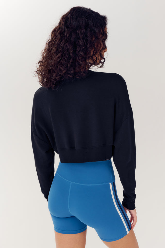 A woman from behind, wearing a SPLITS59 Noah Fleece Crop Sweatshirt in Black and blue athletic shorts, standing against a plain background.