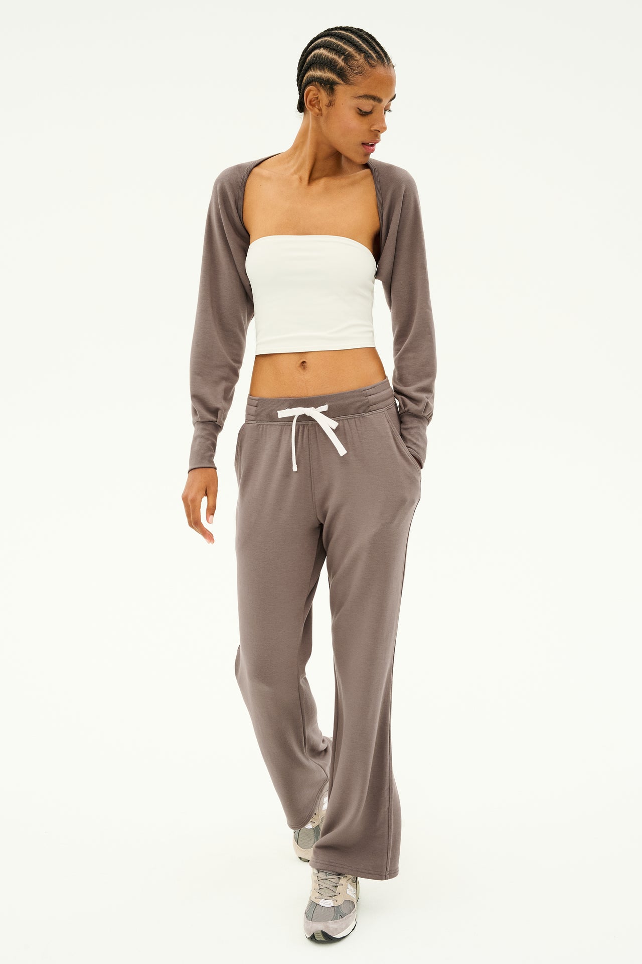 A person dressed in a white tube top, brown cardigan, and matching brown pants with sneakers stands against a plain background. This outfit is ideal for dance warmups or layering under the SPLITS59 Sue Fleece Shrug in Lentil.