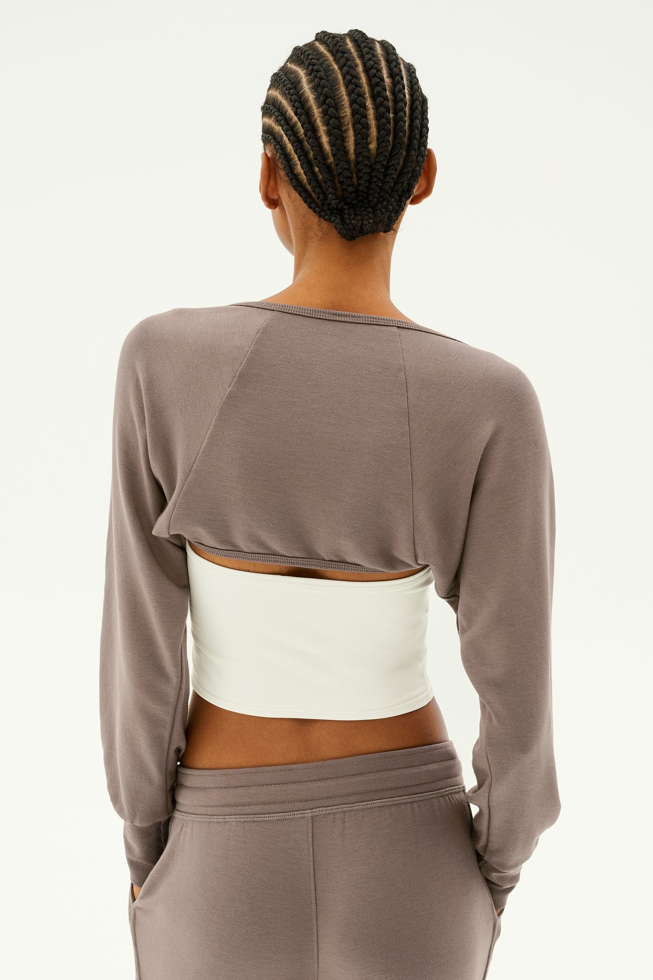 A person with a cornrow hairstyle is seen from behind against a plain background, wearing the SPLITS59 Sue Fleece Shrug in Lentil—a taupe, backless sweatshirt styled over a white top, reminiscent of dance warmups.