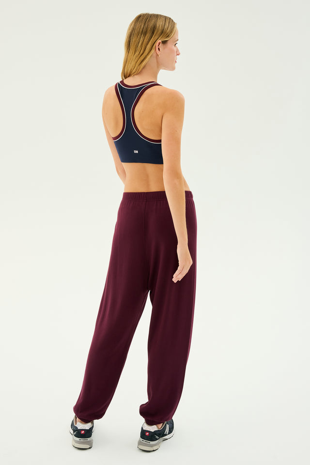 A person with long blonde hair is standing and facing away, wearing a navy sports bra, SPLITS59's Andie Oversized Fleece Sweatpant in Dark Cherry, perfect for cool weather workouts, and black sneakers.