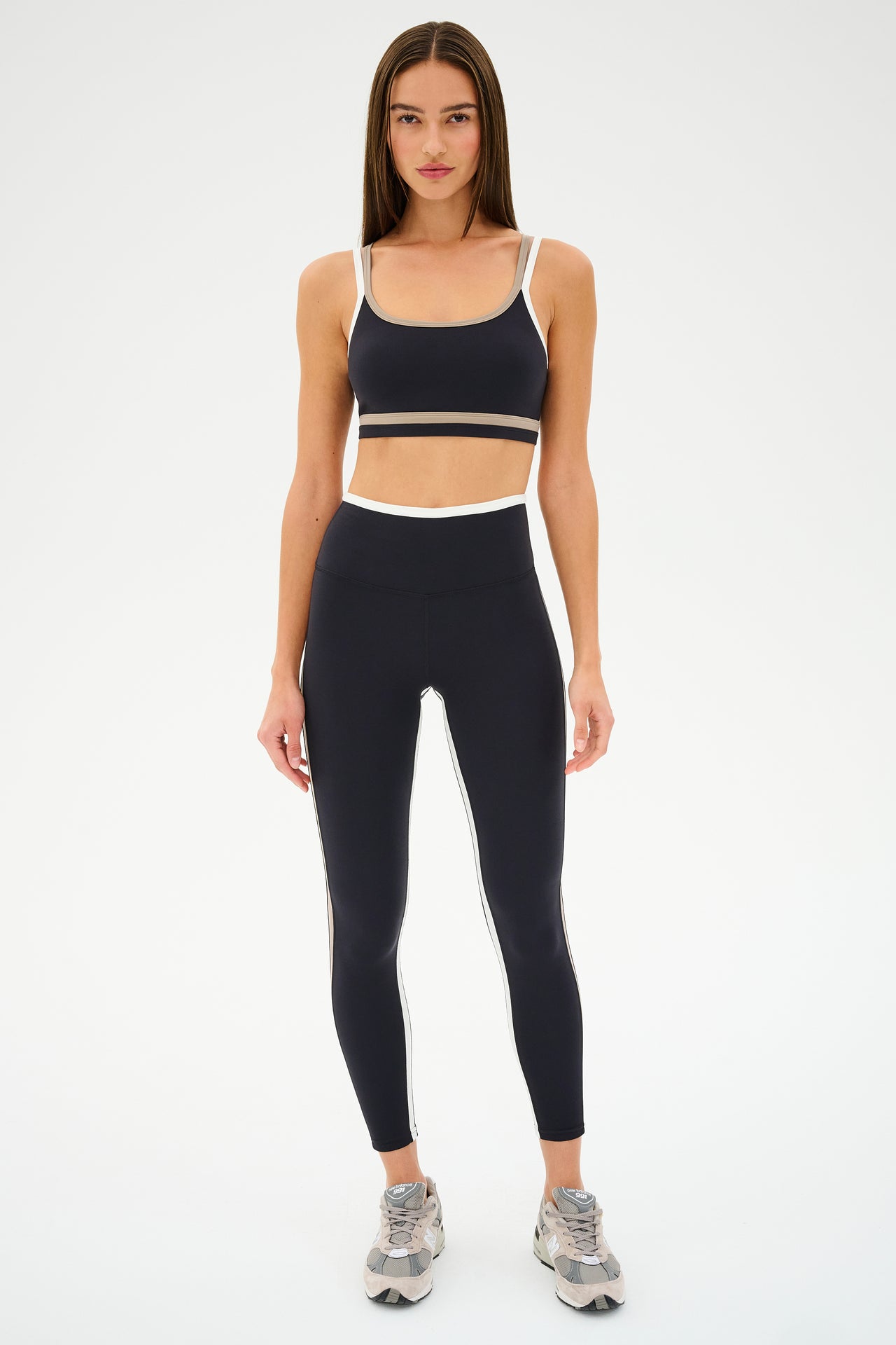 Against a plain background, someone confidently wears the SPLITS59 Eva Rigor Double Strap Bra in Black/Latte, paired with sleek black leggings featuring white accents.