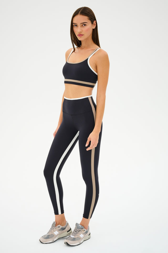 Wearing black and white athletic attire, an individual highlights contrast color details and sneakers against a plain backdrop, completing the look with a SPLITS59 Eva Rigor Double Strap Bra in Black/Latte for added style.