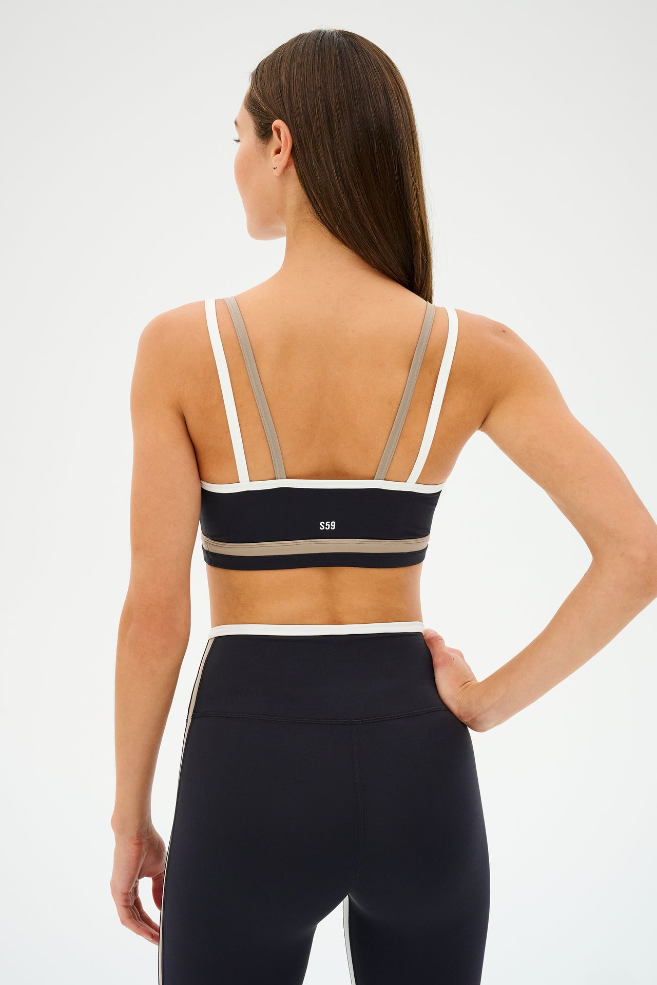 A person in workout attire stands with their back to the camera, wearing a SPLITS59 Eva Rigor Double Strap Bra in Black/Latte, paired with high-waisted leggings.