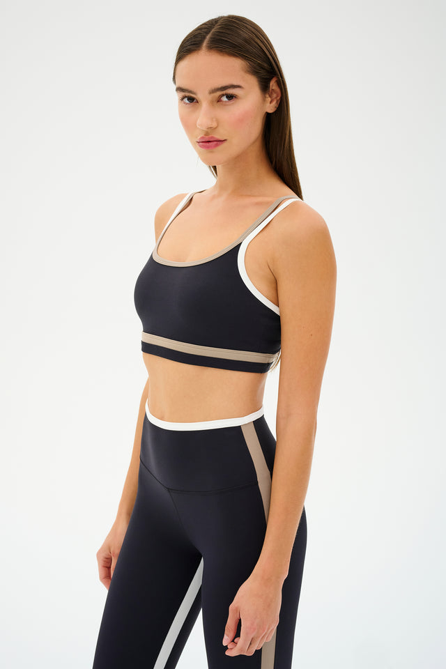 A woman models the SPLITS59 Eva Rigor Double Strap Bra in Black/Latte with matching leggings, highlighting its perfect fit for gym workouts against a plain backdrop.