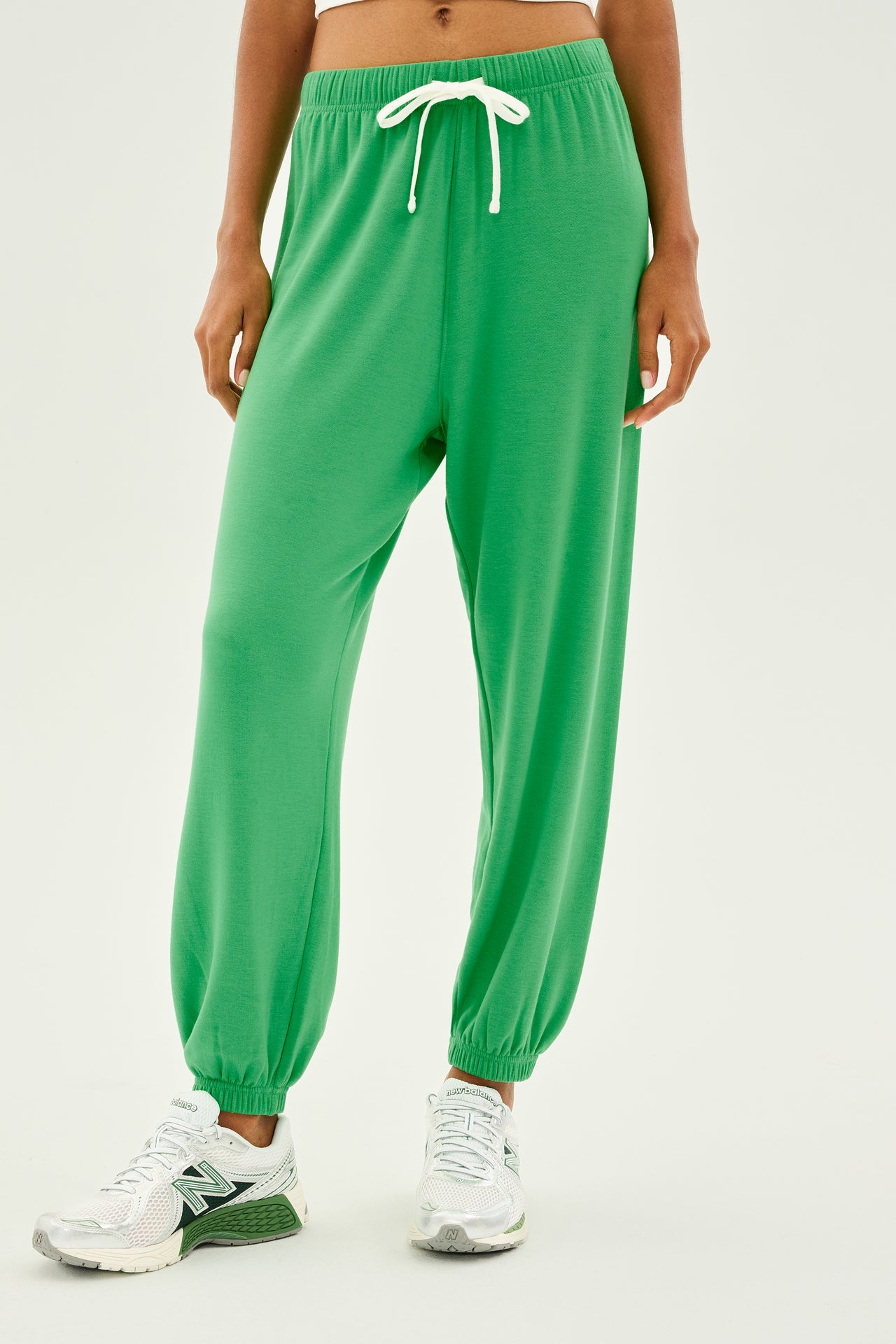 The individual is standing in a neutral pose, wearing the Andie Oversized Fleece Sweatpant in a vibrant grass color from SPLITS59, complete with a drawstring. These stylish sweatpants are paired with crisp white sneakers. Designed and crafted in Los Angeles, they seamlessly combine comfort and style.