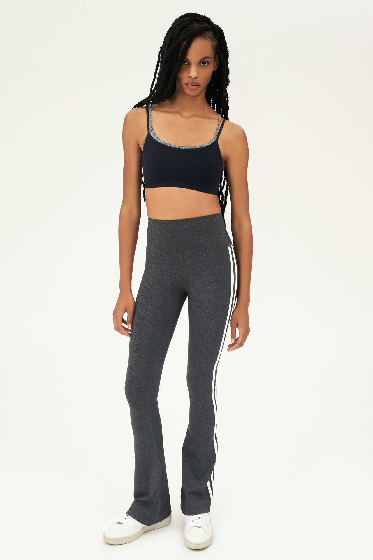Full front view of woman with black braided hair wearing dark gray high waist below ankle length legging with wide flared bottoms and double white stripes on both legs. Also double dark gray and black racerback bra with spaghetti straps. Paired with white shoes.