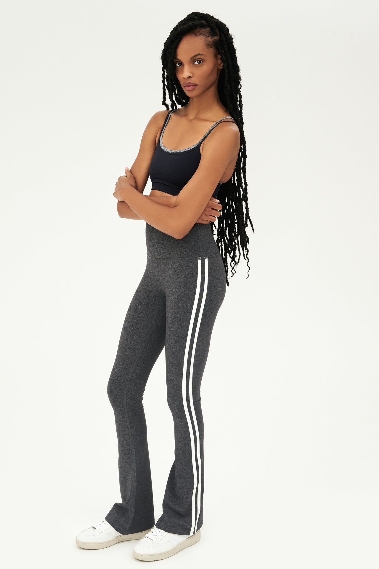 Full front side view of woman with black braided hair wearing dark gray high waist below ankle length legging with wide flared bottoms and double white stripes on both legs. Also double dark gray and black racerback bra with spaghetti straps. Paired with white shoes.